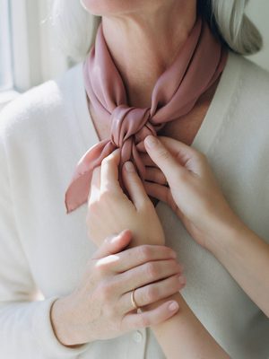 Hands tying a dusty rose silk scarf over a cream knit sweater